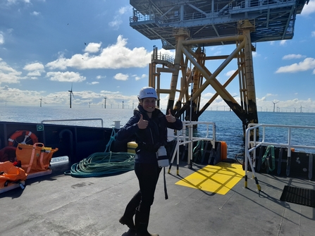 Alice smiling with her thumbs up, stood on the deck of a boat near to the offshore wind substation.